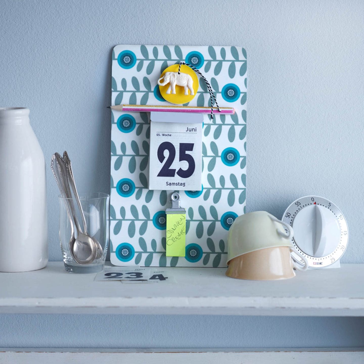 Ein Kalender aus einem gemusterten Brettchen. Ein Minikalender und eine Klammer für ein Notizblock sind daran befestigt