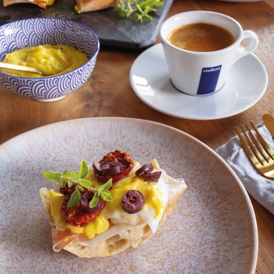 Ciabatta mit Ei, getrockneten Tomaten und Oliven belegt auf altrosa Keramik. Dazu Kaffee (Lavazza) und Dip.