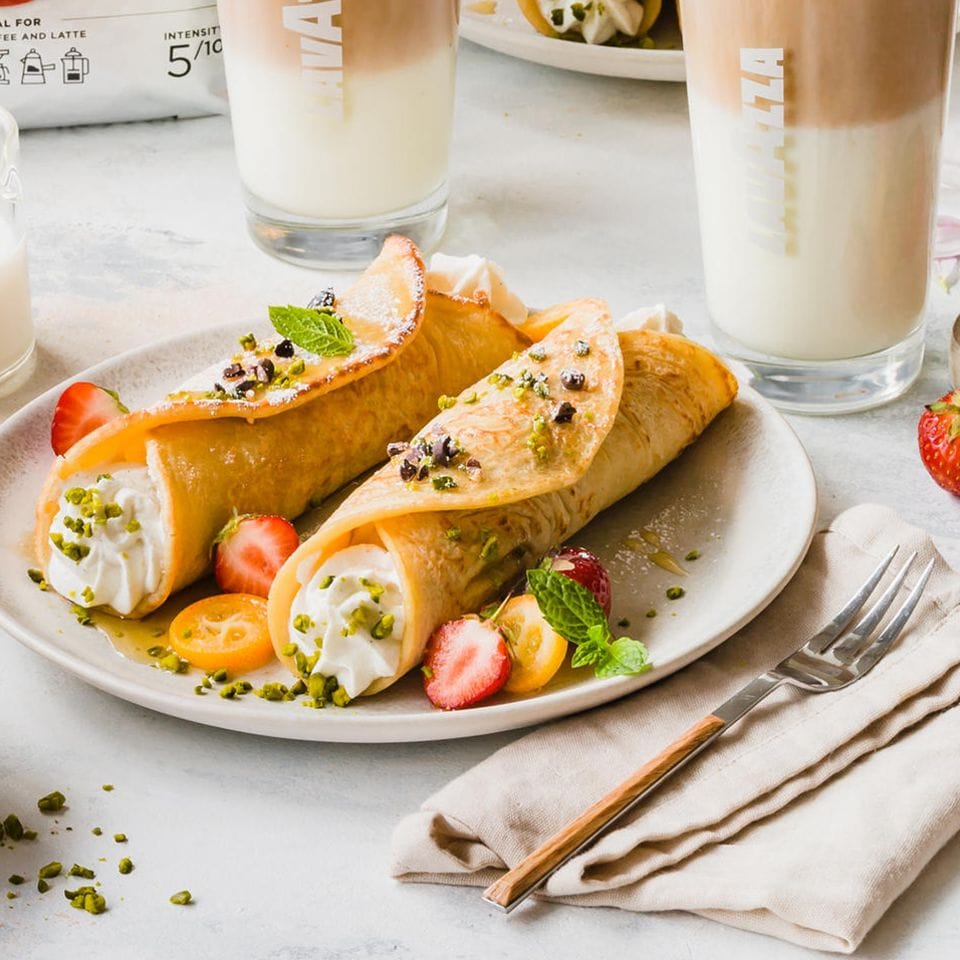 Zwei gerollte Pancakes mit Füllung neben mediterraner Tischdeko und Latte Macchiato