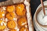 Gebackener Blechkuchen belegt mit Orangenscheiben und Mandeln steht neben Schüsseln mit geschlagener Sahne und Karamellsauce