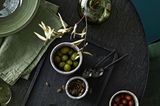 Auf einem schwarzen Holztisch steht ein Tablett mit Oliven & eine grüne Glasvase, daneben liegen verschiedene Textilien in Oliv