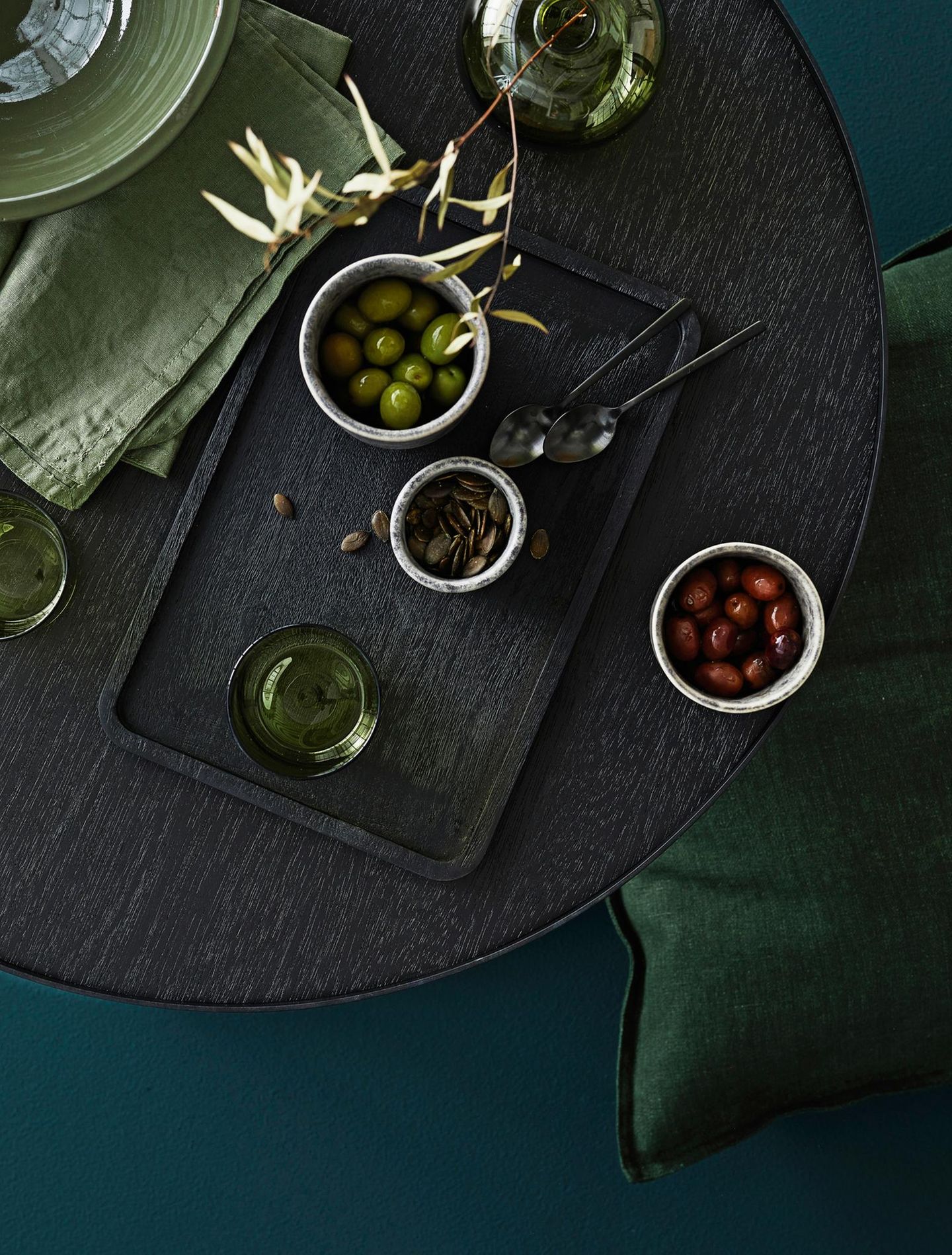 Auf einem schwarzen Holztisch steht ein Tablett mit Oliven & eine grüne Glasvase, daneben liegen verschiedene Textilien in Oliv