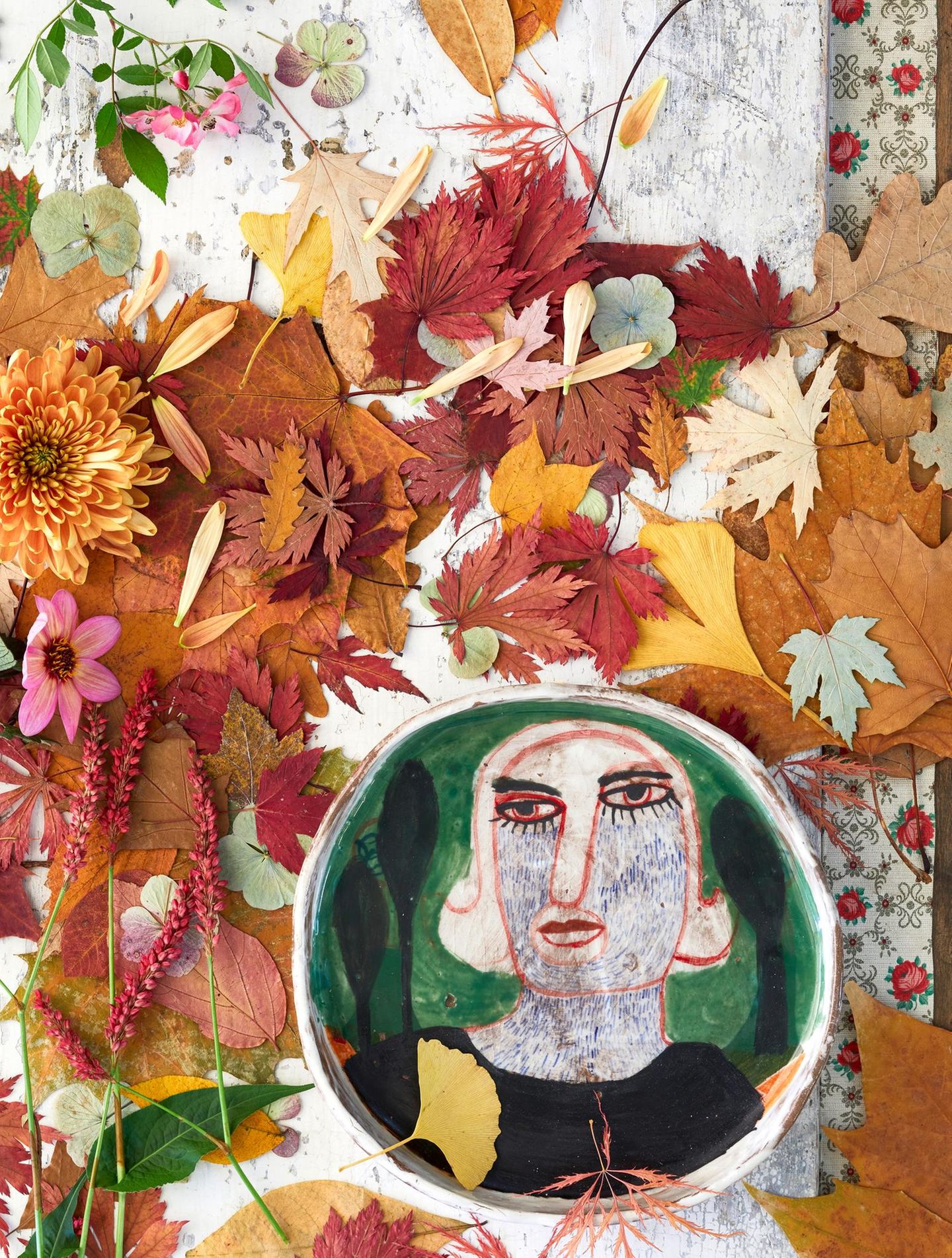 Ein Keramikteller mit abstrakter Darstellung eines Menschen steht auf einem Tisch mit Laub und Herbstblumen