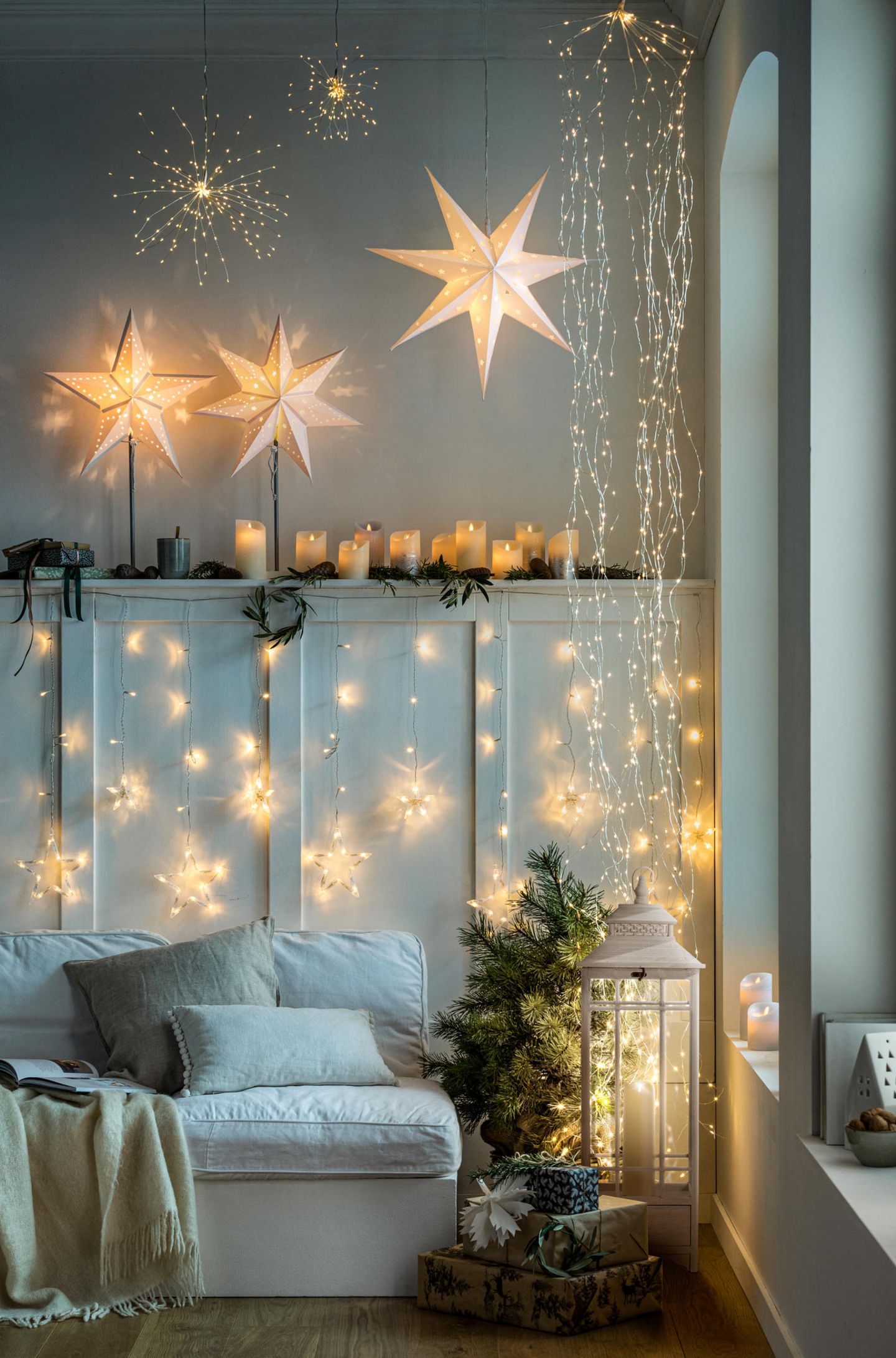 Lichterketten an den Wänden und leuchtende Sterne, die von der Decke hängen, in einem Wohnzimmer mit Sofa und kleiner Tanne