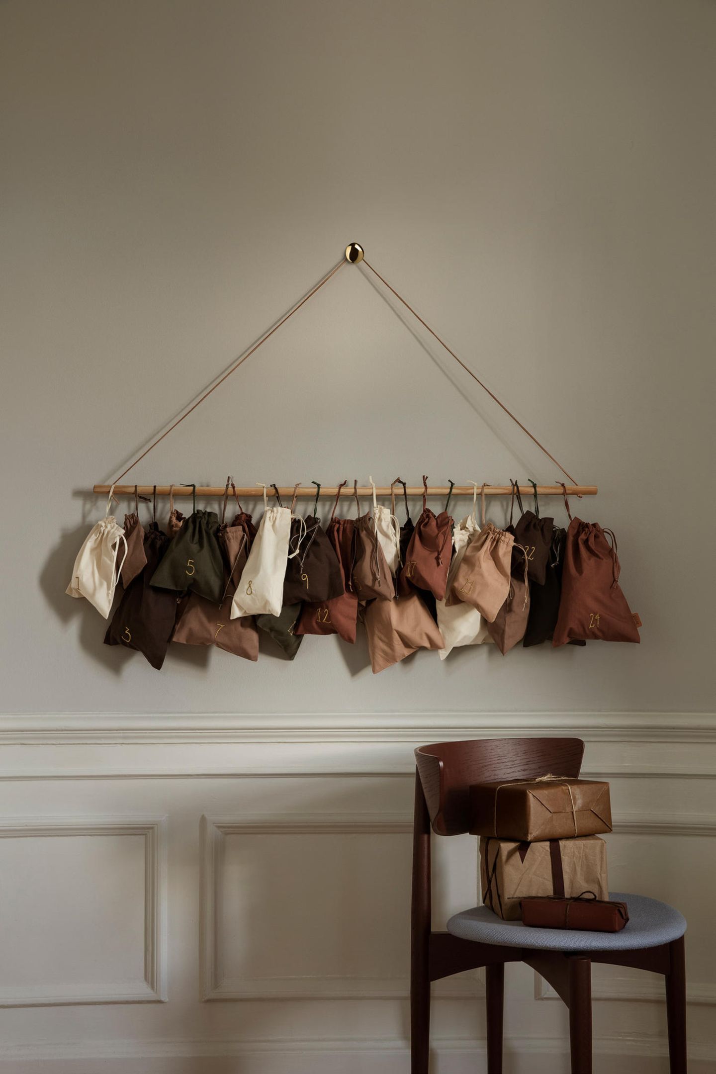 Adventskalender mit kleinen Säcken in verschiedenen Brauntönen hängen an einem Stock an einer Wand; darunter ein dunkler Stuhl