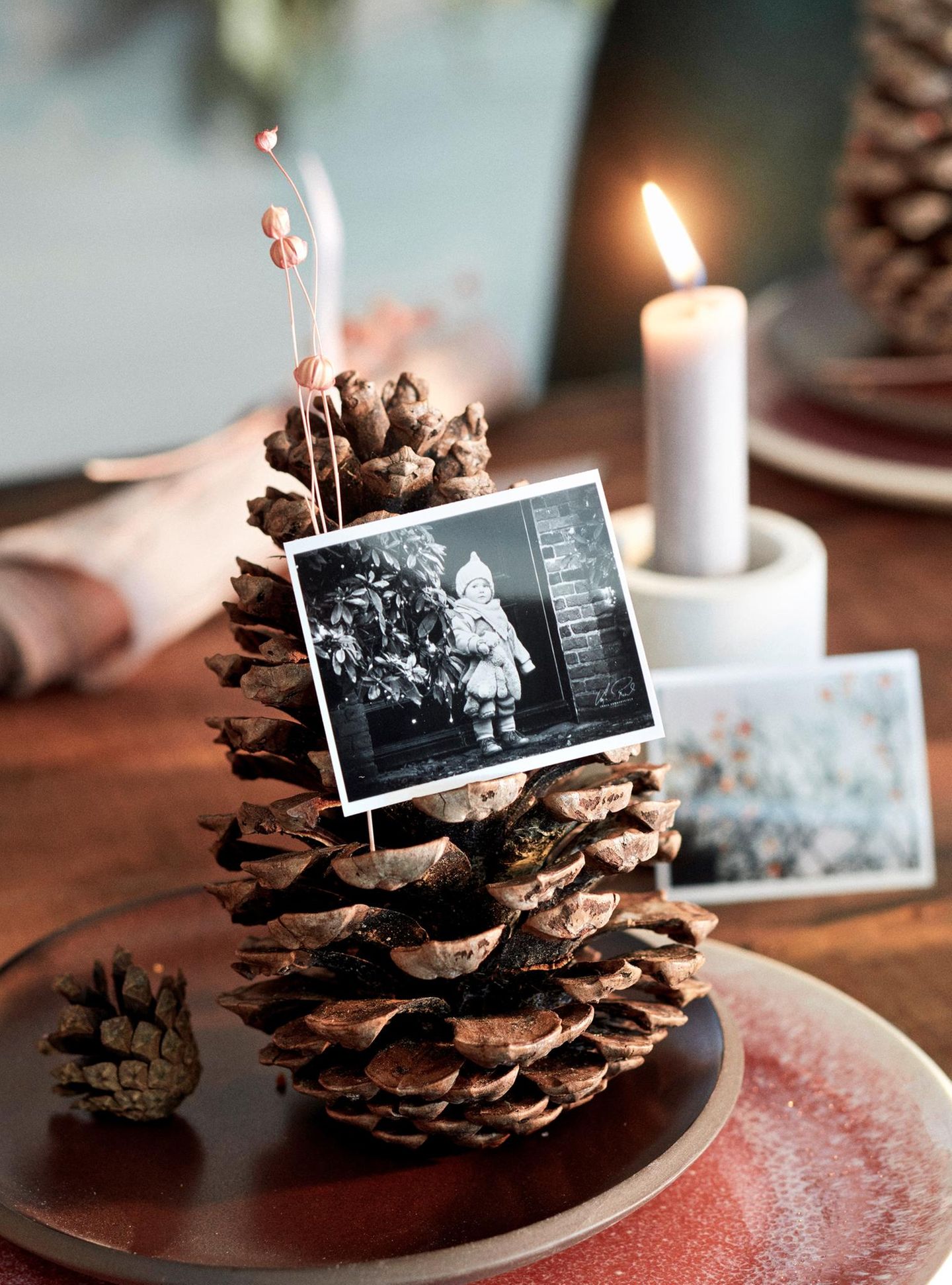 Ein Foto ist in einen Tannenzapfen geklemmt, der auf einem braunen Keramikteller steht; im Hintergrund steht eine Kerze