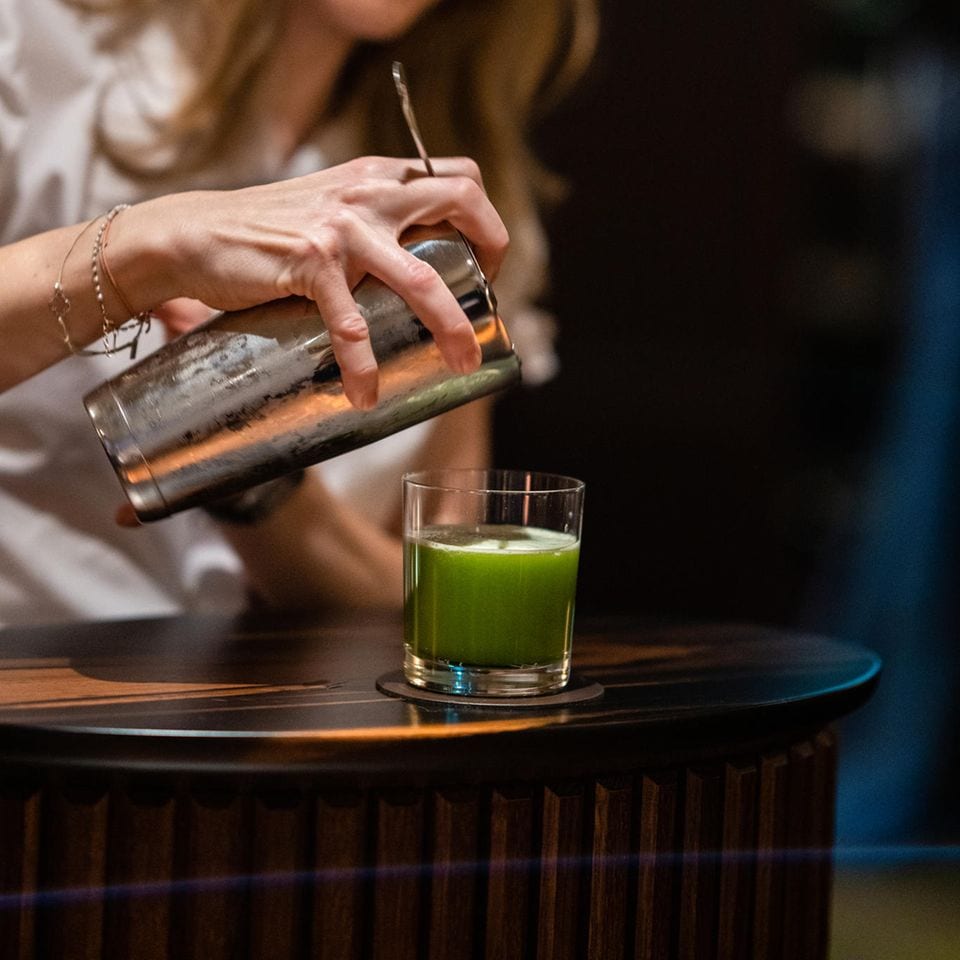 Frau gießt aus einem silbernen Cocktail-Shaker einen grünen Drink in ein Glas auf einem Holztisch