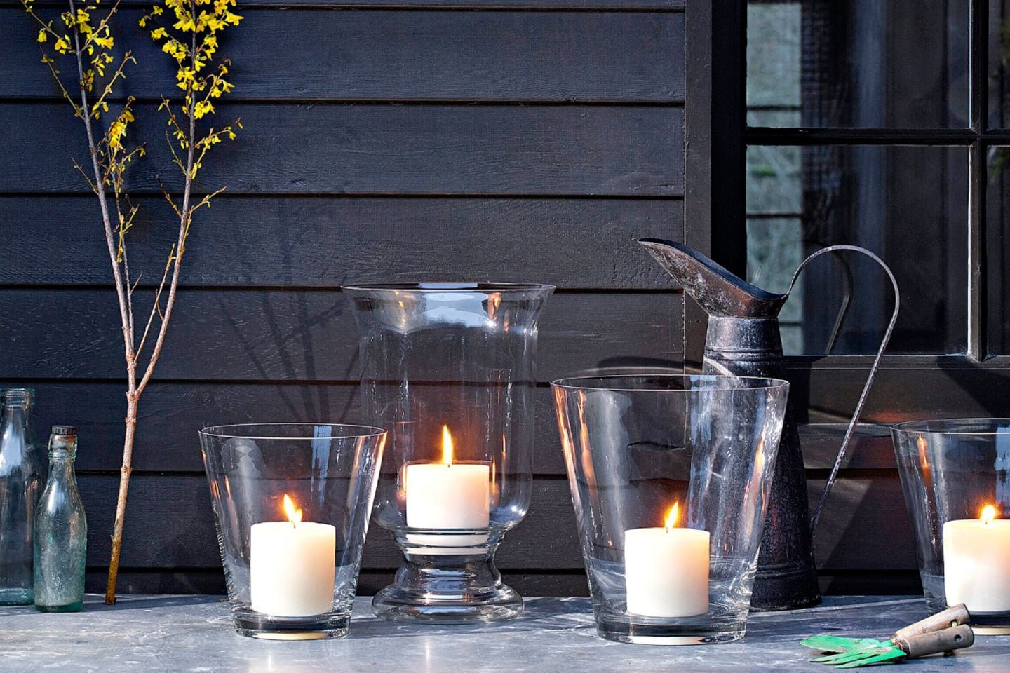 Gartentisch mit vier Kerzen in Vasen als Windlichter vor einer hölzernen Hauswand