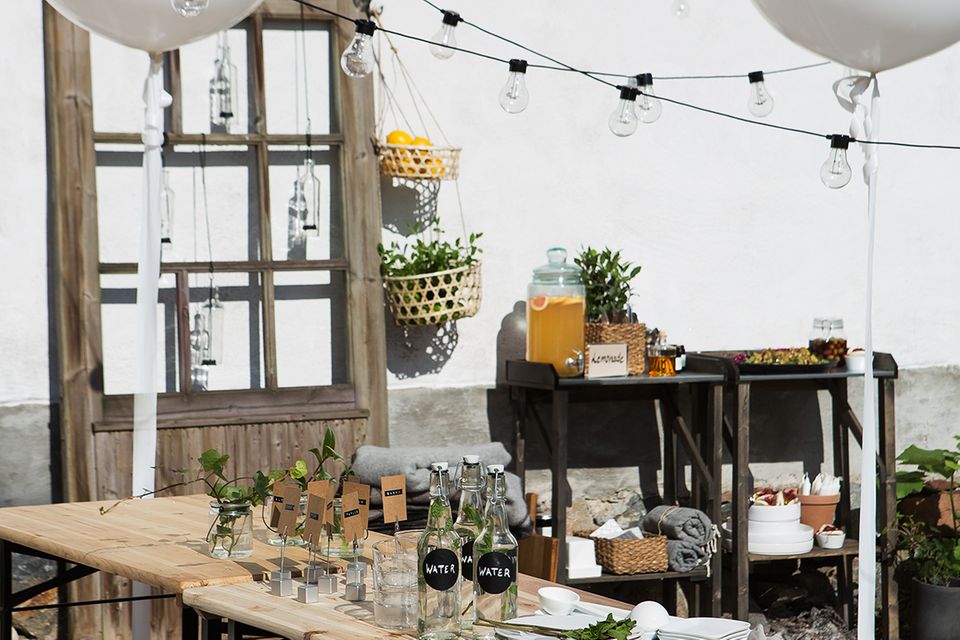 Mit Gläsern und Tellern gedeckte Tische auf einer Terrasse, darüber baumeln Lichterketten und weiße Ballons