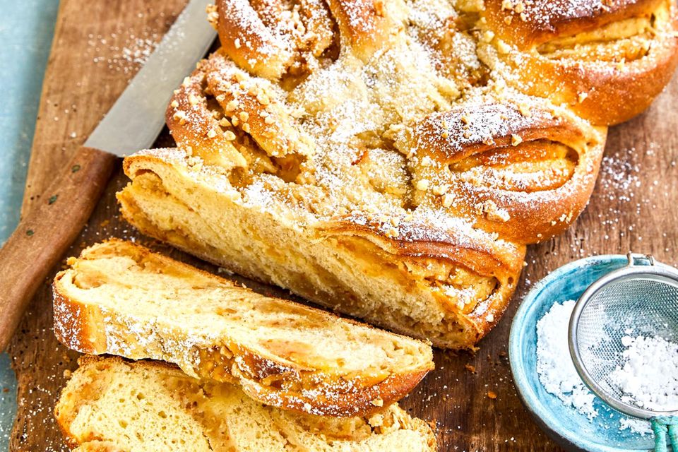 Der Osterzopf mit Füllung ist mit Puderzucker bestreut und  liegt auf einem braunen Brett.