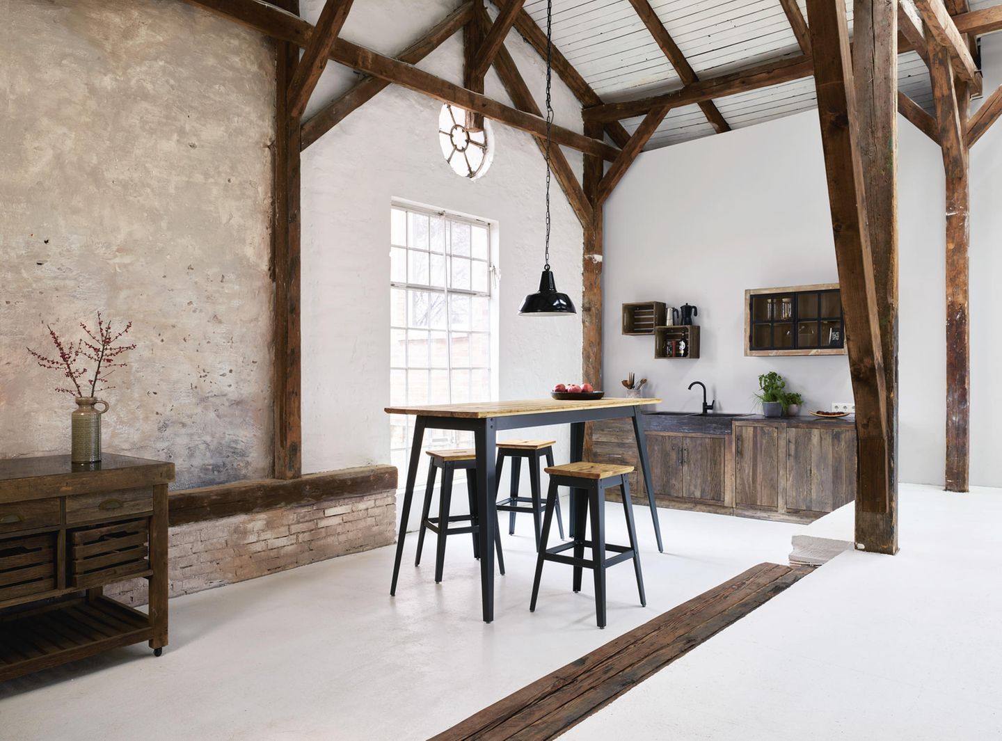 Eine dunkle Küche aus Holz mit Betonboden, dunklen Holzträgern, in der Mitte steht ein hoher Esstisch mit Hockern