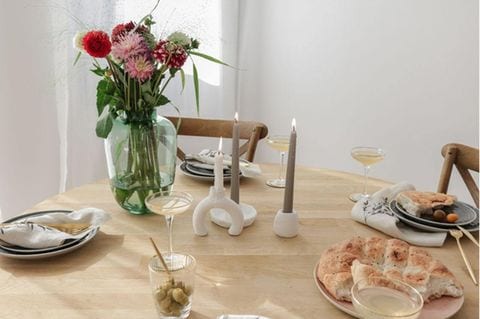 Ein gedeckter Holztisch mit Blumen in einer Vase, drei selbstgemachten Kerzenständern sowie Brot und Getränken