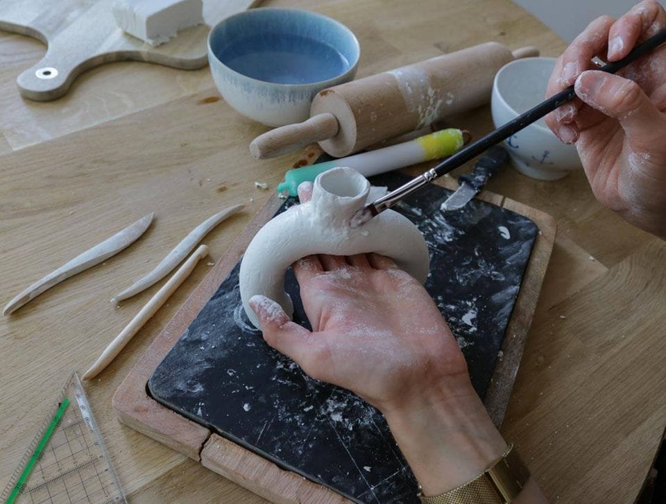Ein Kerzenständer aus Ton wird mit einem Pinsel bestrichen, daneben ein Nudelholz, Keramikschalen und Schneidebretter