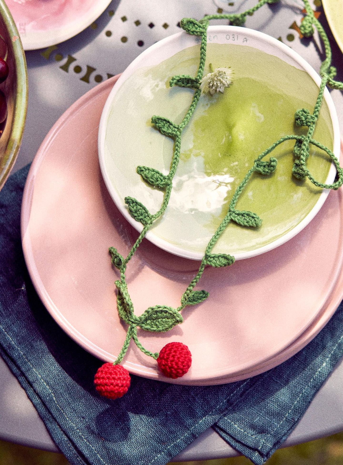 Eine gehäkelte Kette mit Kirschmotiven liegt auf pinken und grünen Tellern, darunter eine blaue Serviette