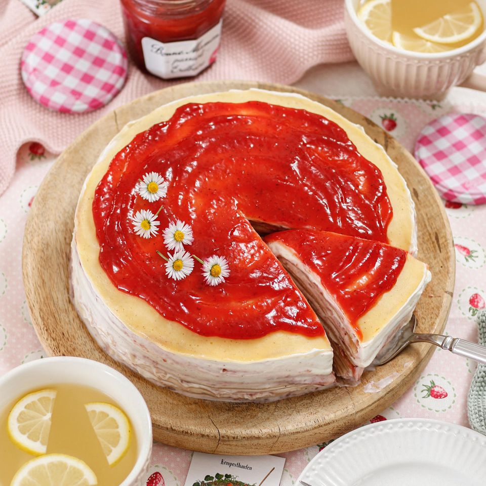Eine Torte aus Crêpes mit Erdbeer-Creme und einer Schicht Marmelade steht auf einem gedeckten Kaffeetisch