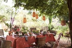 Großer Tisch im Freien mit roter Leinentischdecke, Geschirr, Rattanlampen an einem Baum und Stühlen