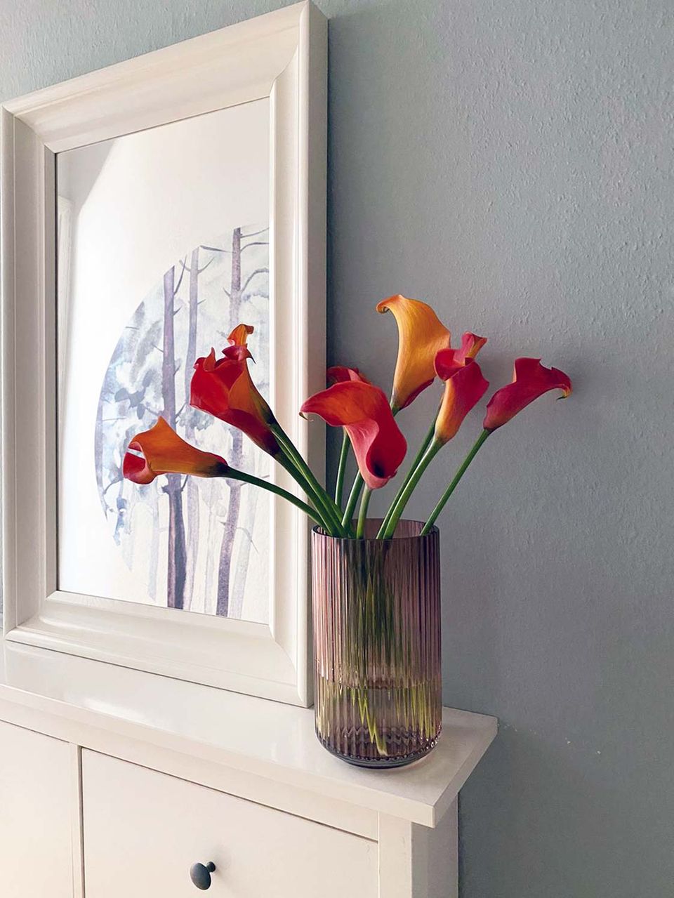Glasvase mit roten und orangenen Blumen auf einer weißen Kommode