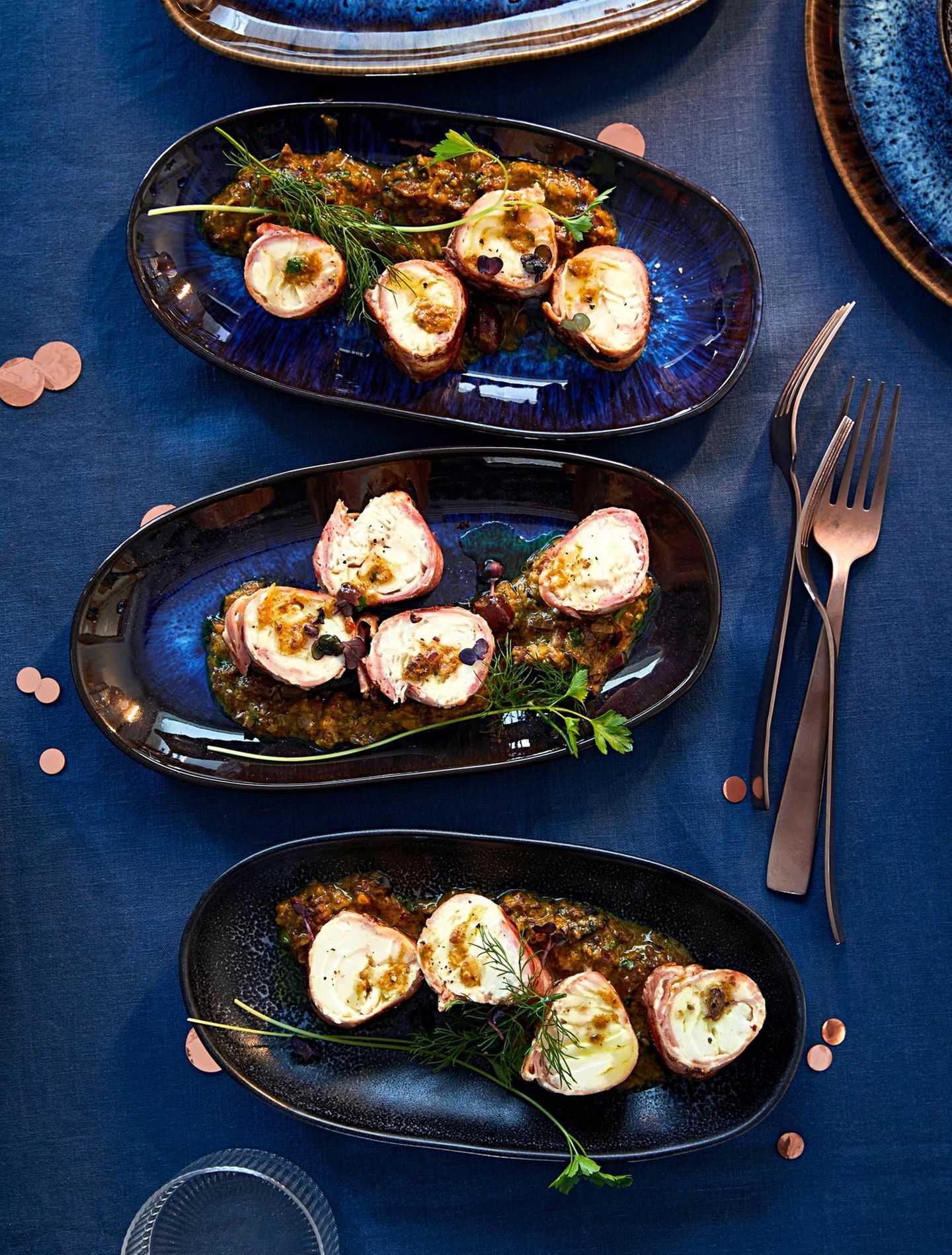 Röllchen aus Kabeljau mit Bacon und Kräutern auf blauen Tellern angerichtet