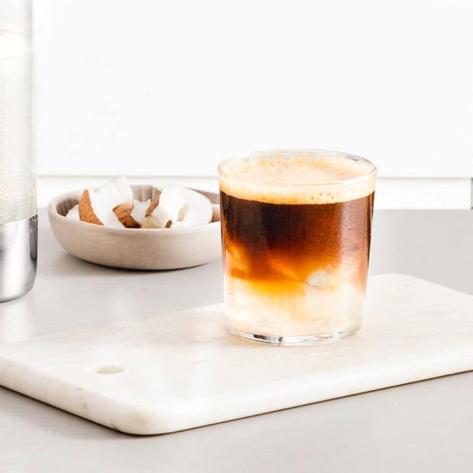 glas mit kaffee und eis auf einem weißen brett, dahinter kokosnusssscheiben in einer schale