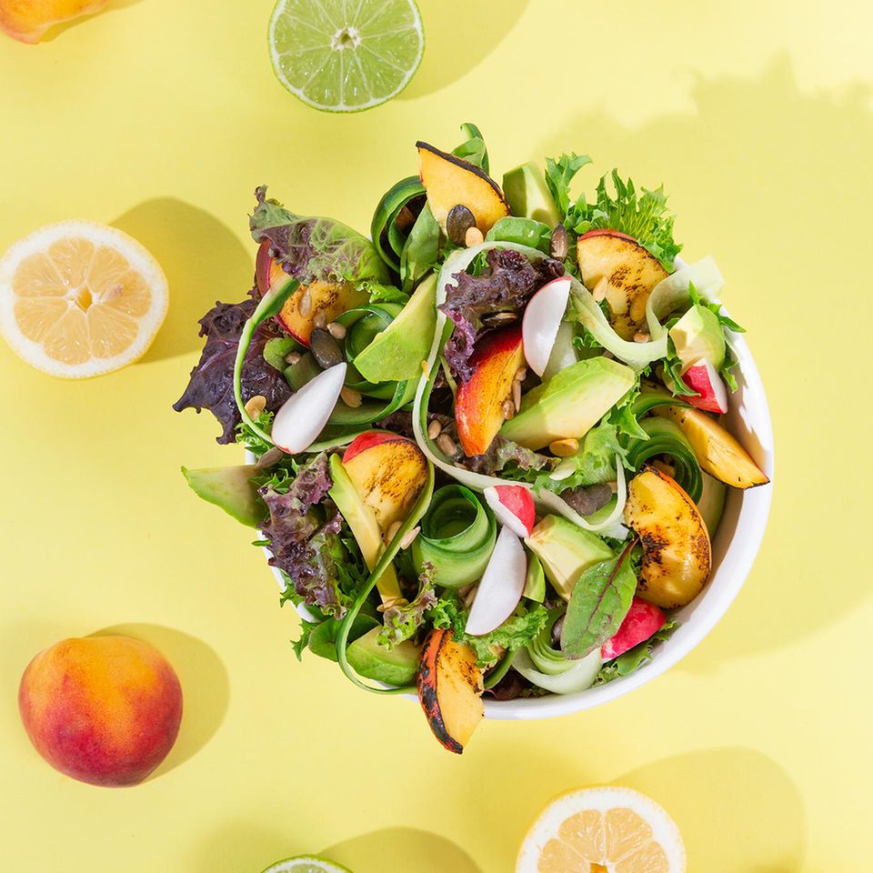 Salat mit Mango und Pfirsich und Avocado in einer Schüssel, dahinter gelber Hintergrund und Früchte verteilt