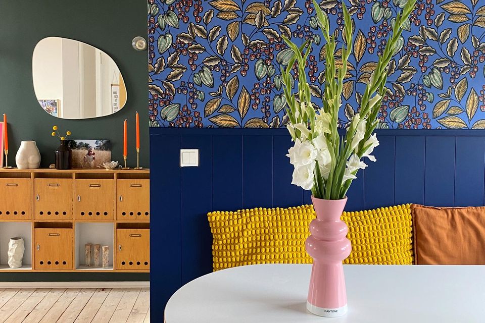Bunte Tapete mit dunkelblauer Holzverkleidung, davor ein runder Tisch mit pinker Vase und Blumen sowie gelben Kissen
